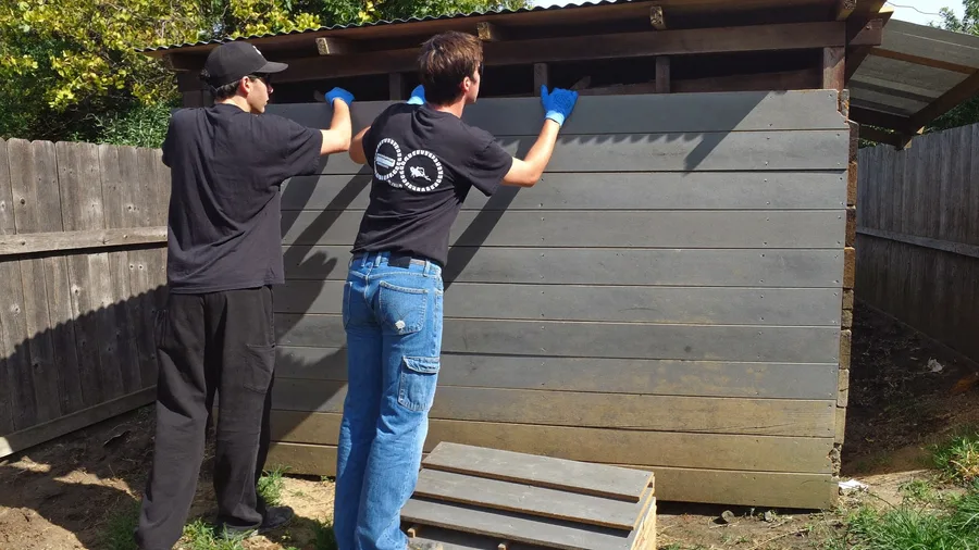 Two workers tearing down an old wooden garden shed in a backyard